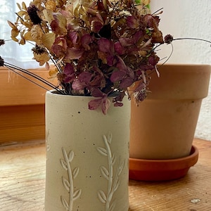 Pu&ograve; includere: Un vaso in ceramica beige con un design botanico, contenente fiori secchi nei toni del viola, marrone e beige. Un vaso di terracotta &egrave; sullo sfondo. Il vaso ha una forma cilindrica con motivi a foglie in rilievo.