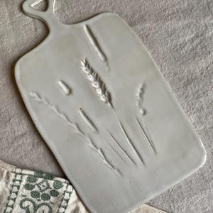 Könnte beinhalten: Ein rechteckiges, cremeweißes Keramik-Servierbrett mit einem Griff mit Aussparung. Die Oberfläche ist mit einem detaillierten botanischen Muster aus Weizenähren und Gräsern geprägt und weist eine glatte, glasierte Oberfläche mit feinen dunklen Sprenkeln auf.