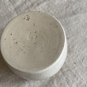 May include: A circular ceramic cup base in an off-white speckled finish. The bottom surface features a textured, matte appearance with scattered dark brown flecks and a small, stamped maker's mark consisting of a stylized symbol near the edge.