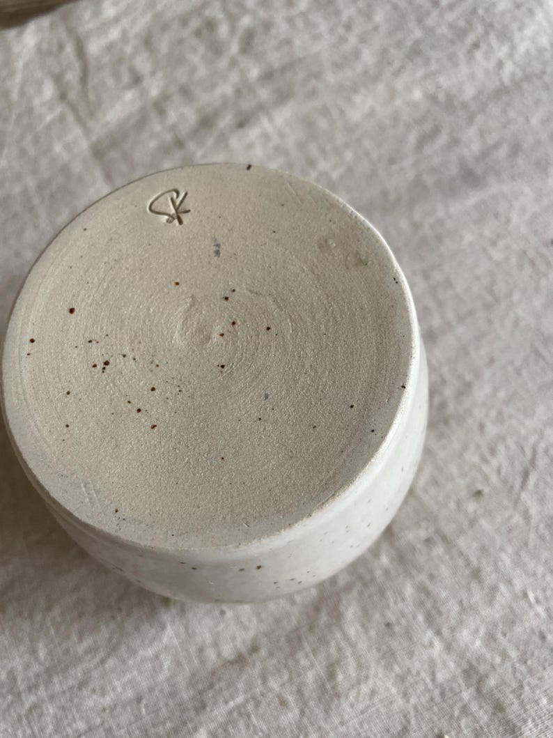 May include: A circular ceramic cup base in an off-white speckled finish. The bottom surface features a textured, matte appearance with scattered dark brown flecks and a small, stamped maker's mark consisting of a stylized symbol near the edge.