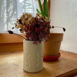 Pu&ograve; includere: Un piccolo vaso in ceramica bianco sporco con un motivo a foglie in rilievo, contenente fiori secchi nei toni del marrone e del bordeaux. Un vaso di terracotta con una pianta &egrave; appoggiato su un davanzale di legno, con una tenda trasparente sullo sfondo.