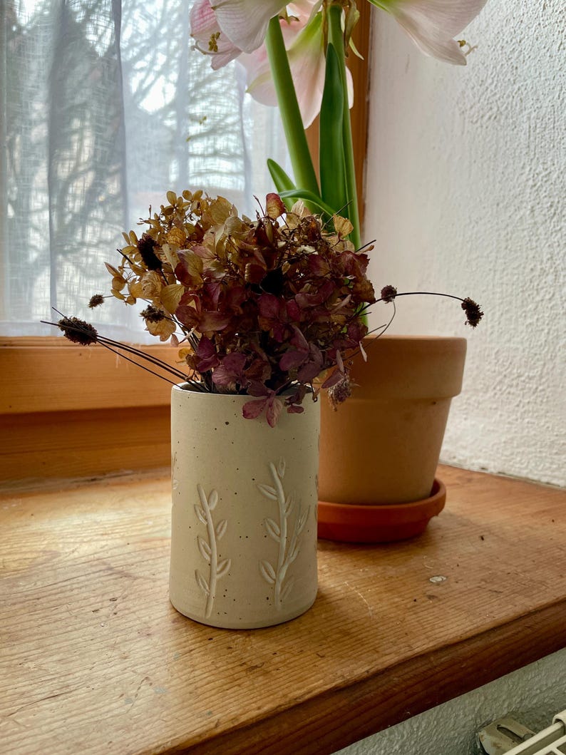 Pu&ograve; includere: Un piccolo vaso in ceramica bianco sporco con un motivo a foglie in rilievo, contenente fiori secchi nei toni del marrone e del bordeaux. Un vaso di terracotta con una pianta &egrave; appoggiato su un davanzale di legno, con una tenda trasparente sullo sfondo.