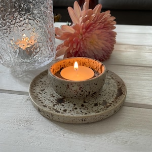 May include: A lit tealight candle in a speckled ceramic bowl and saucer. The bowl and saucer are beige with brown speckles. A textured glass vase and a pink flower are in the background.