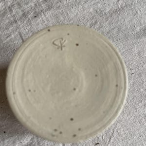 May include: A circular ceramic base with a speckled off-white glaze and a smooth, flat surface. A small, hand-stamped maker's mark featuring a stylized symbol is clearly visible near the top edge of the clay surface, showing subtle texture.