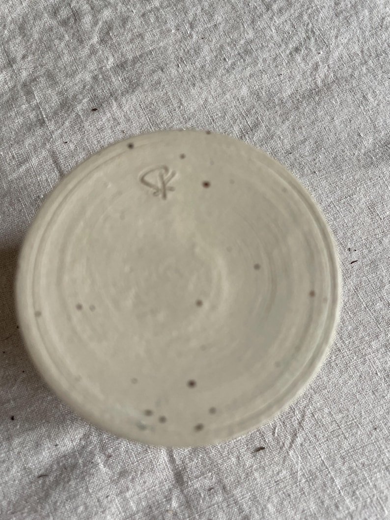 May include: A circular ceramic base with a speckled off-white glaze and a smooth, flat surface. A small, hand-stamped maker's mark featuring a stylized symbol is clearly visible near the top edge of the clay surface, showing subtle texture.