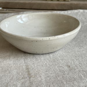 Könnte beinhalten: Eine runde, flache Keramikschale mit einer glänzenden, cremeweißen Glasur und sichtbaren dunkelbraunen Sprenkeln auf der gesamten Oberfläche. Die Schale hat ein glattes, geschwungenes Inneres und eine leicht konisch zulaufende Außenwand.