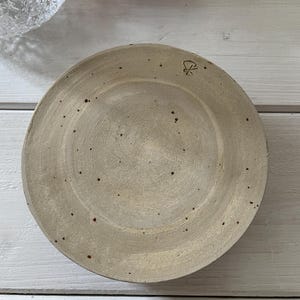 May include: A round, beige ceramic plate with a slightly raised rim. The plate has a speckled appearance with small, dark brown spots. A small, stylized symbol is visible on the upper right side of the plate.