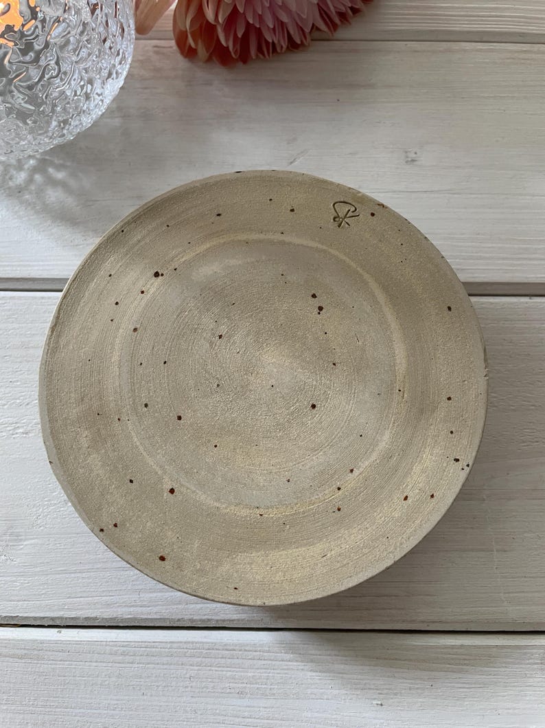 May include: A round, beige ceramic plate with a slightly raised rim. The plate has a speckled appearance with small, dark brown spots. A small, stylized symbol is visible on the upper right side of the plate.