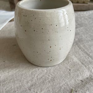 May include: A barrel-shaped ceramic cup with a smooth, off-white glaze and a speckled finish featuring small dark brown dots scattered across the exterior surface. The vessel has a wide, open top and a rounded body, showcasing a handcrafted appearance.