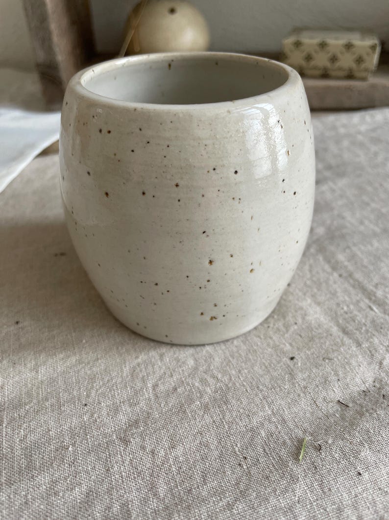 May include: A barrel-shaped ceramic cup with a smooth, off-white glaze and a speckled finish featuring small dark brown dots scattered across the exterior surface. The vessel has a wide, open top and a rounded body, showcasing a handcrafted appearance.