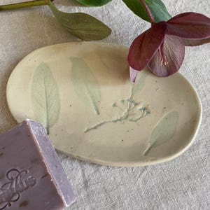May include: An oval ceramic soap dish in a light cream colour featuring embossed green leaf and botanical imprints. A rectangular bar of lavender-coloured soap with embossed text is positioned at the bottom left corner of the dish.