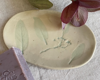 May include: An oval ceramic soap dish in a light cream color featuring embossed green leaf and botanical imprints. A rectangular bar of lavender-colored soap with embossed text is positioned at the bottom left corner of the dish.