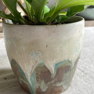 May include: A ceramic plant pot featuring a smooth, off-white upper section that transitions into a textured, dripped glaze pattern of earthy brown and muted teal tones. A vibrant green leafy plant grows from the top of the cylindrical container.