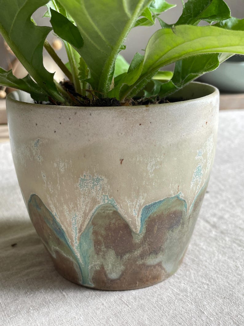 May include: A ceramic plant pot featuring a smooth, off-white upper section that transitions into a textured, dripped glaze pattern of earthy brown and muted teal tones. A vibrant green leafy plant grows from the top of the cylindrical container.
