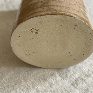 May include: A cylindrical ceramic vessel with a light beige base featuring dark speckles and a smooth, glossy tan glaze on the upper portion. The bottom surface displays a small, incised maker's mark consisting of a stylized heart and cross symbol.