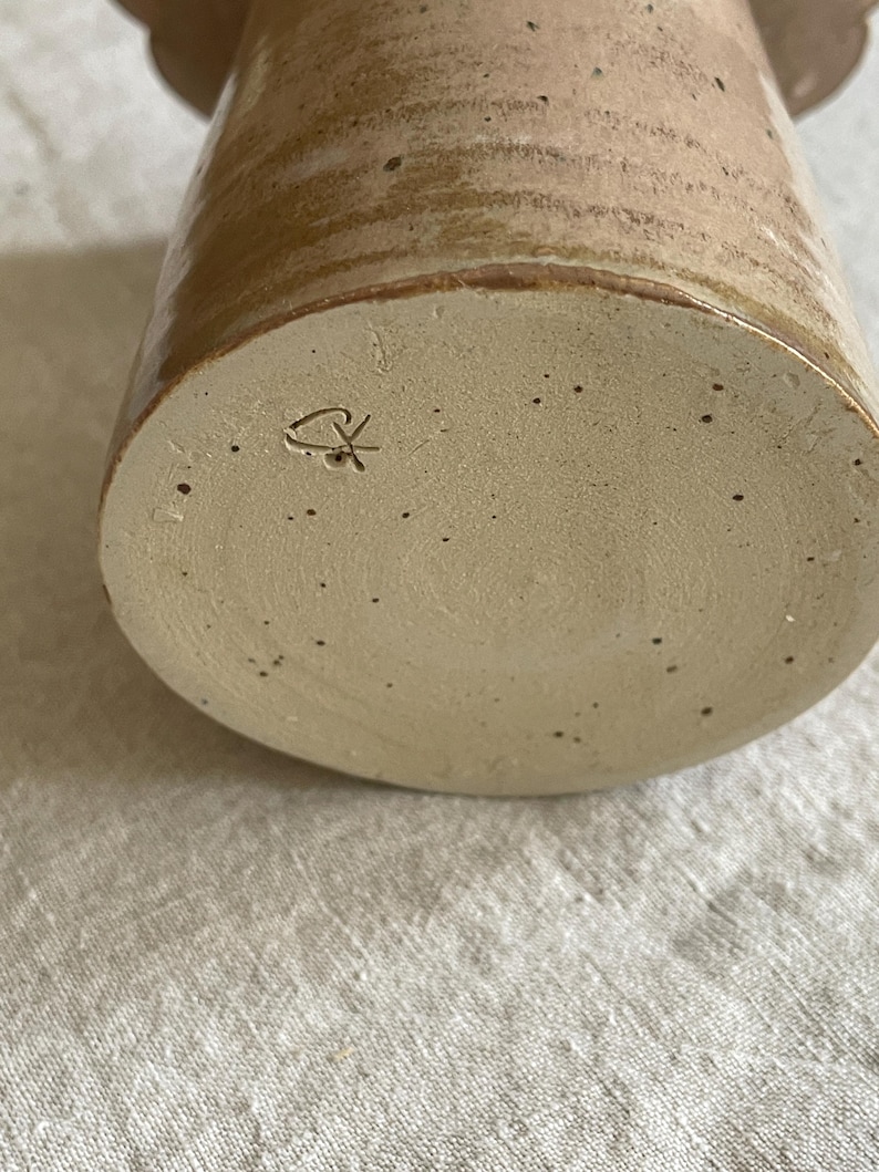 May include: A cylindrical ceramic vessel with a light beige base featuring dark speckles and a smooth, glossy tan glaze on the upper portion. The bottom surface displays a small, incised maker's mark consisting of a stylized heart and cross symbol.