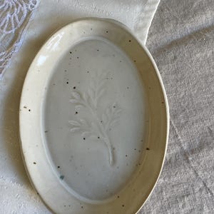 May include: An oval ceramic dish with a glossy off-white glaze and a speckled finish. The centre of the dish features a subtle, embossed botanical leaf design. The edges are slightly raised, creating a shallow basin for holding small items.