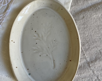 Può includere: Un piatto ovale in ceramica con una lucida smaltatura bianco sporco e una finitura maculata. Il centro del piatto presenta un sottile disegno botanico a foglia in rilievo. I bordi sono leggermente rialzati, creando una conca poco profonda.