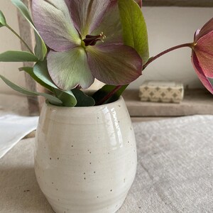 May include: A small, off-white ceramic vase with a speckled finish containing a bouquet of purple and green hellebore flowers and eucalyptus leaves. The vase has a rounded, bulbous shape with a smooth, glossy glaze and subtle horizontal ridges.