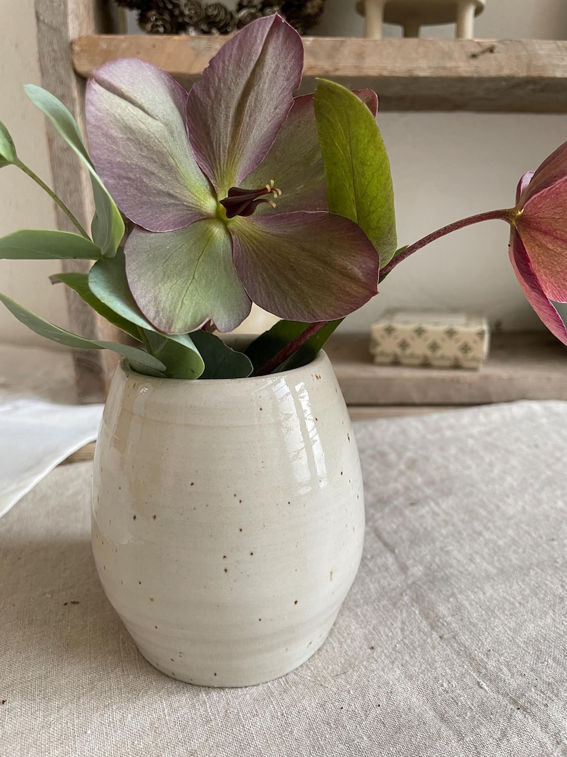 May include: A small, off-white ceramic vase with a speckled finish containing a bouquet of purple and green hellebore flowers and eucalyptus leaves. The vase has a rounded, bulbous shape with a smooth, glossy glaze and subtle horizontal ridges.