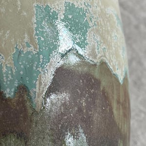 May include: A ceramic vase with a glossy, multi-layered glaze featuring a smooth transition between cream, turquoise, and dark brown tones. The surface displays a reactive, mottled texture where the colors overlap, creating a layered effect.
