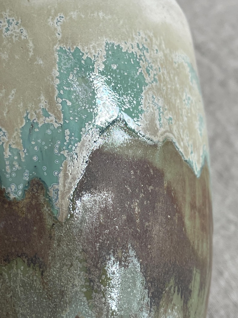 May include: A ceramic vase with a glossy, multi-layered glaze featuring a smooth transition between cream, turquoise, and dark brown tones. The surface displays a reactive, mottled texture where the colors overlap, creating a layered effect.