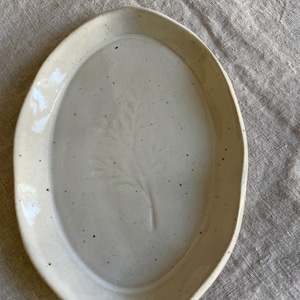 May include: An oval-shaped ceramic dish with a glossy off-white glaze and a speckled finish. The centre of the dish features a subtle, embossed botanical leaf pattern. The edges of the dish are slightly raised, creating a shallow basin.