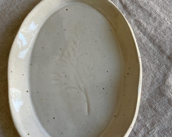 Può includere: Un piatto in ceramica di forma ovale con una finitura lucida color bianco sporco e un aspetto maculato. Il centro del piatto presenta un sottile motivo botanico a foglia in rilievo. I bordi del piatto sono leggermente rialzati.