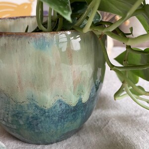 May include: A round ceramic planter with a glossy glaze featuring a gradient of deep teal at the base transitioning into a pale mint green and soft pink toward the rim. A green leafy plant with variegated foliage grows from the interior.