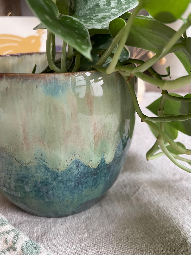May include: A round ceramic planter with a glossy glaze featuring a gradient of deep teal at the base transitioning into a pale mint green and soft pink toward the rim. A green leafy plant with variegated foliage grows from the interior.