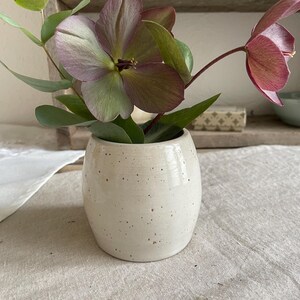 May include: A small, off-white ceramic vase with a speckled finish containing a bouquet of hellebore flowers in shades of dusty purple and green, accompanied by sprigs of eucalyptus leaves with rounded foliage.
