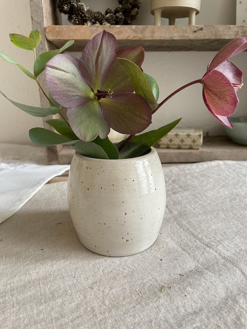May include: A small, off-white ceramic vase with a speckled finish containing a bouquet of hellebore flowers in shades of dusty purple and green, accompanied by sprigs of eucalyptus leaves with rounded foliage.
