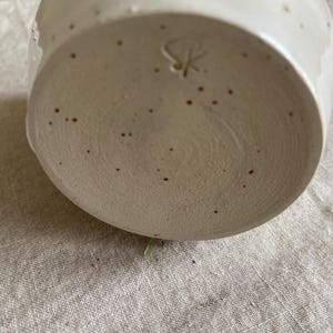 May include: A ceramic mug base with a speckled off-white glaze and a textured, unglazed bottom surface. The base features a hand-carved maker's mark consisting of the letters S and K intertwined, centered on the circular bottom of the vessel.