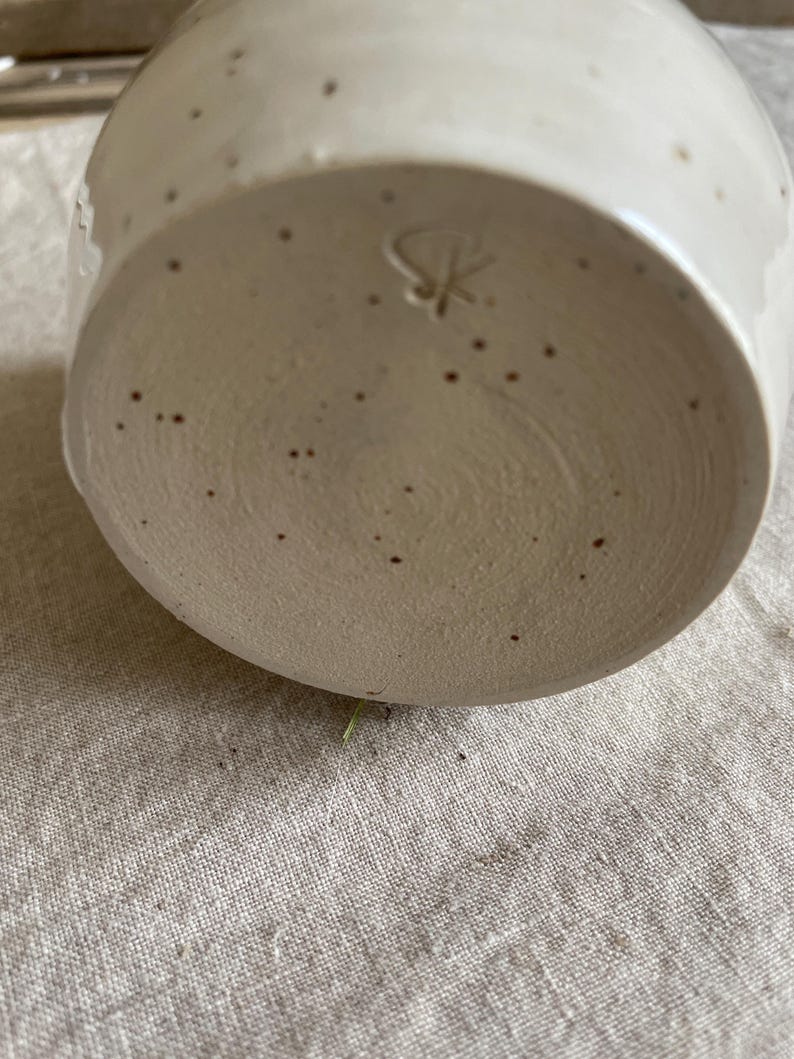 May include: A ceramic mug base with a speckled off-white glaze and a textured, unglazed bottom surface. The base features a hand-carved maker's mark consisting of the letters S and K intertwined, centered on the circular bottom of the vessel.