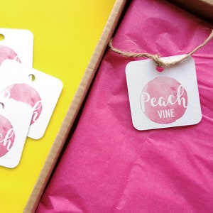 Puede incluir: Una caja de regalo rosa con una etiqueta blanca que dice "Peach Vine" en un c&iacute;rculo rosa de acuarela. La etiqueta est&aacute; atada con un cordel y la caja est&aacute; llena de papel de seda rosa.