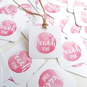 Puede incluir: Un mont&oacute;n de etiquetas de regalo cuadradas blancas con un c&iacute;rculo de acuarela rosa y el texto "Peach Vine" en blanco. Las etiquetas est&aacute;n esparcidas sobre una superficie blanca.