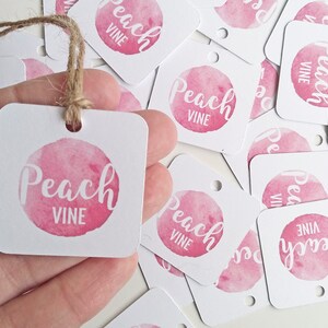 Puede incluir: Una pila de etiquetas de regalo cuadradas blancas con un c&iacute;rculo de acuarela rosa y el texto "Peach Vine" en blanco. Las etiquetas tienen un agujero perforado en la esquina superior y est&aacute;n atadas con un cordel.