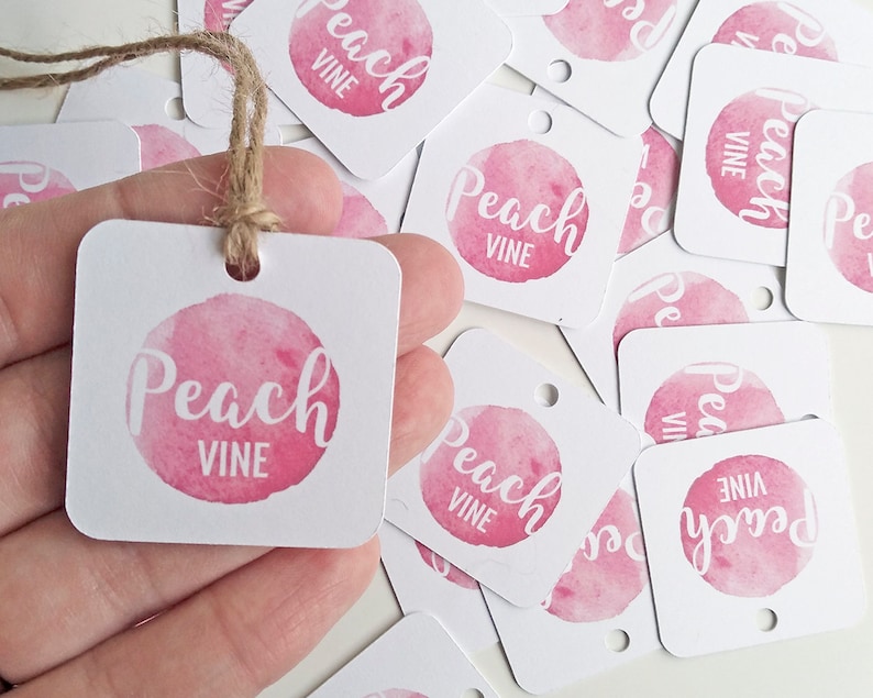 Puede incluir: Una pila de etiquetas de regalo cuadradas blancas con un c&iacute;rculo de acuarela rosa y el texto "Peach Vine" en blanco. Las etiquetas tienen un agujero perforado en la esquina superior y est&aacute;n atadas con un cordel.