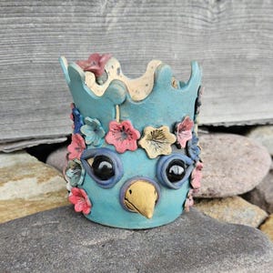 May include: A light blue ceramic cup featuring a sculpted bird face with large black eyes and a yellow beak. The exterior is decorated with various small, colourful clay flowers in shades of pink, blue, and yellow. The top rim has a wavy edge.