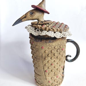 May include: A quirky ceramic mug featuring a bird-like sculpture. The mug is tan with red dots and a dark handle. The bird has a long beak, a small red hat, and a decorative collar. A unique, handcrafted piece of pottery.