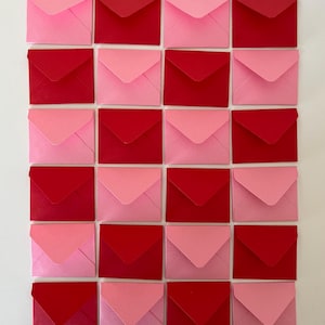 Puede incluir: Una cuadrícula de veinticuatro sobres pequeños en tonos rosa y rojo. Los sobres están dispuestos en un patrón cuadrado, alternando entre rosa claro y rojo. Todos los sobres están cerrados y tienen una forma cuadrada clásica.