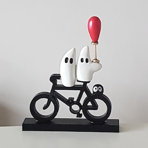 May include: A whimsical wooden sculpture featuring two white, ghost-like figures riding a black bicycle. One figure holds a red balloon. The sculpture is mounted on a black base, creating a playful and artistic display.
