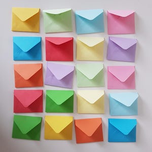 Puede incluir: Una vista cenital muestra una cuadrícula de dieciséis sobres pequeños y cuadrados en varios colores pastel. Los colores incluyen amarillo, verde, azul, rosa, rojo, naranja y morado. Los sobres están dispuestos en cuatro filas de cuatro.