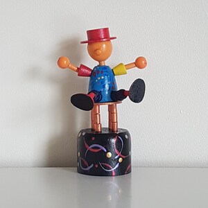 May include: A wooden toy puppet with a red hat, blue shirt, and colourful details. The puppet is on a black and white base with confetti-like designs.