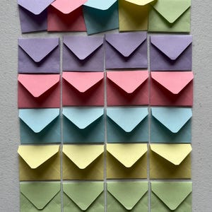 Puede incluir: Una colección de veinticuatro sobres de papel pequeños dispuestos en una cuadrícula. Los sobres son de varios colores pastel, incluyendo morado claro, rosa, azul, amarillo y verde. Cada sobre presenta un cierre de solapa triangular.