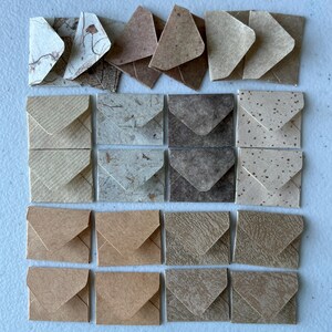 May include: A collection of twenty small square envelopes arranged in a grid. The envelopes feature various textures and shades of brown, tan, beige, and cream, including marbled, speckled, ribbed, and fibrous paper finishes with triangular flaps.