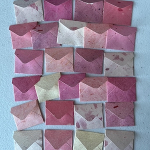 Könnte beinhalten: Eine Sammlung von vierundzwanzig kleinen, quadratischen Umschlägen, die in einem Raster angeordnet sind. Die Umschläge bestehen aus strukturiertem Papier in verschiedenen Rosa-, Magenta-, Creme- und Beigetönen, jeweils mit dreieckiger Klappe und sichtbaren Papierfasern.