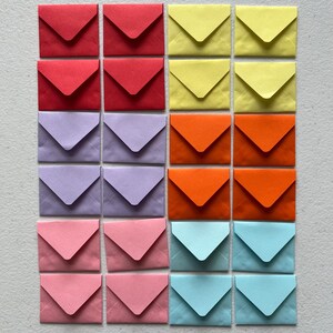 May include: A collection of twenty-four small square envelopes arranged in a grid of four columns and six rows. The envelopes feature a pointed flap and come in a variety of solid colours including red, yellow, light purple, orange, pink, and light blue.