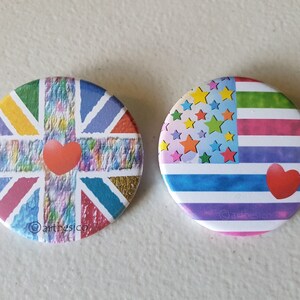 May include: Two round buttons with colourful designs. The first button features a rainbow-coloured Union Jack flag with a red heart in the centre. The second button features a rainbow-coloured American flag with a red heart in the bottom right corner.