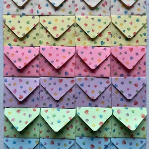 May include: A grid of twenty-four small paper envelopes arranged in six rows and four columns. Each envelope features a repeating pattern of tiny multicolored hearts against a solid pastel background, with colors including cream, yellow, pink, purple, green, and blue.